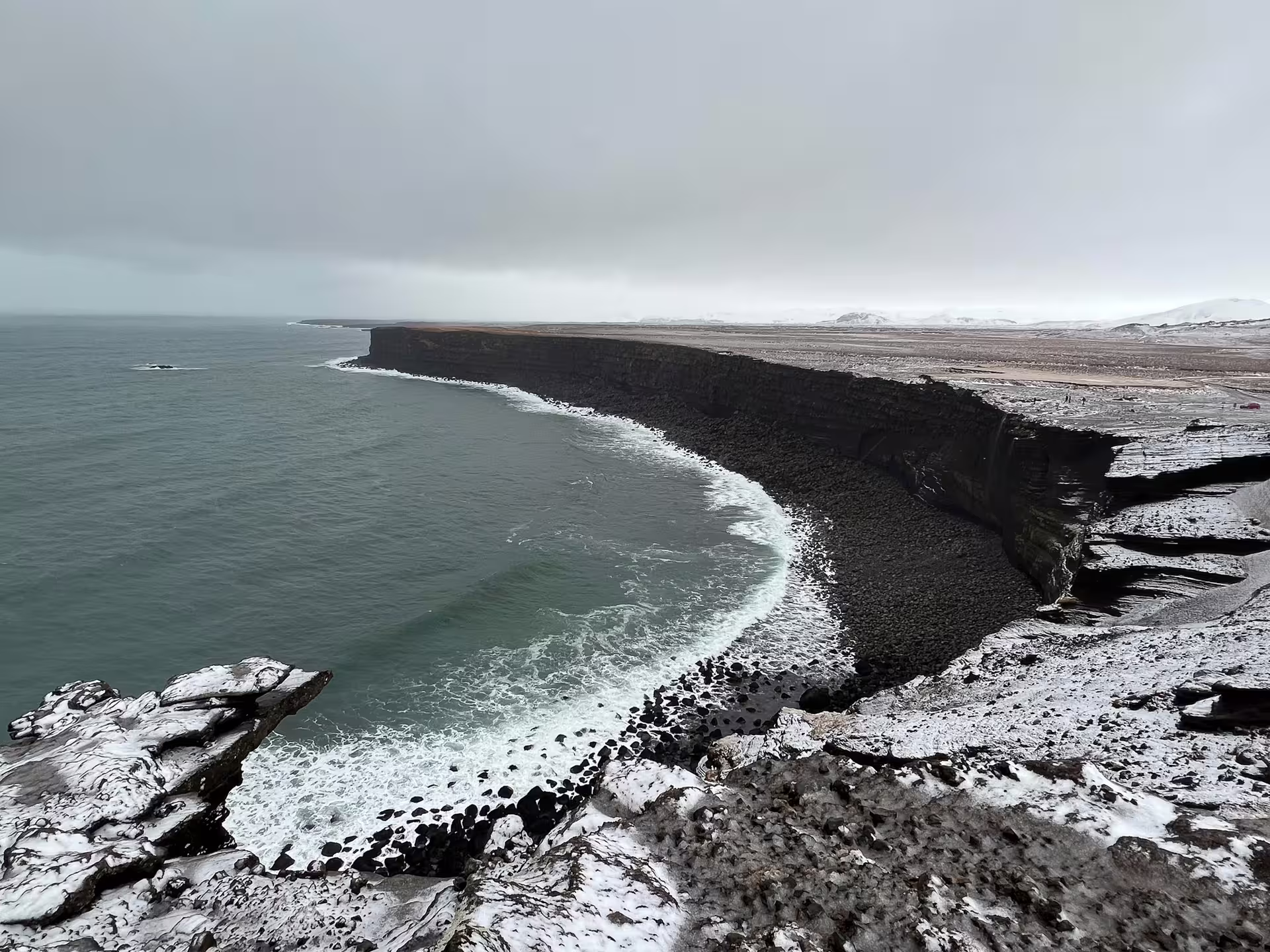 Black volcanic cliffs along Iceland's south coast with snow-dusted rocks and crashing Atlantic waves