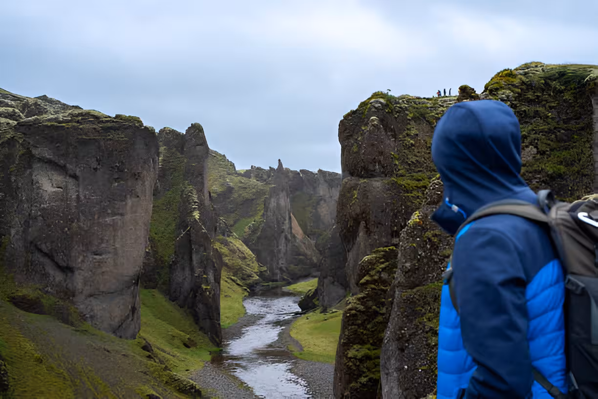 Hiker in blue jacket overlooking mossy Fjaðrárgljúfur canyon with river below on Iceland south coast