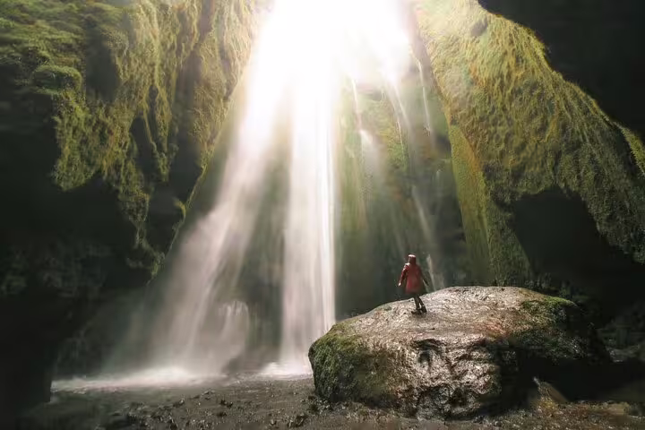 Person in red jacket standing on rock inside cave watching Iceland waterfall with dramatic light rays