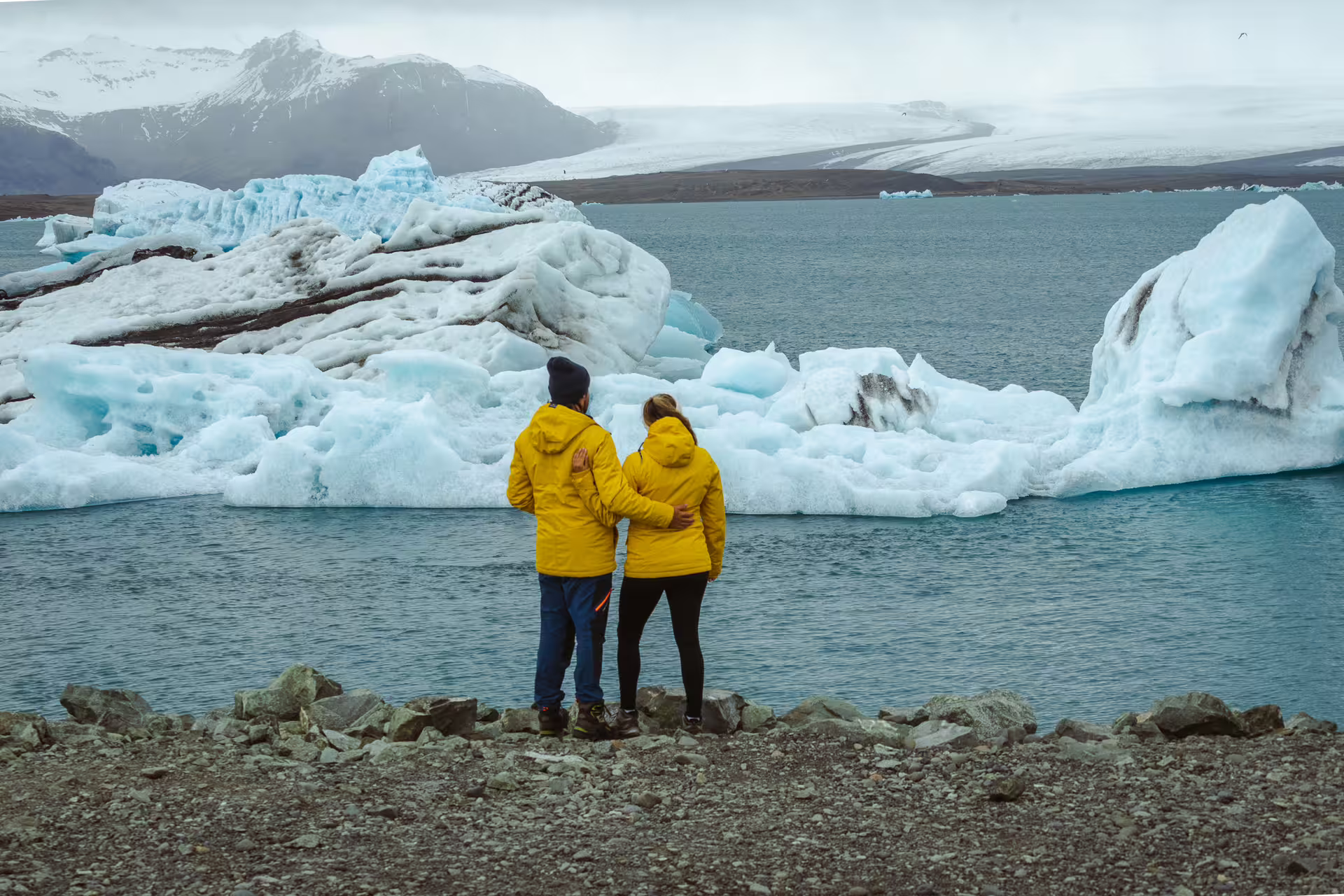 Couple in yellow jackets standing before blue icebergs at Iceland glacier lagoon on South Coast tour