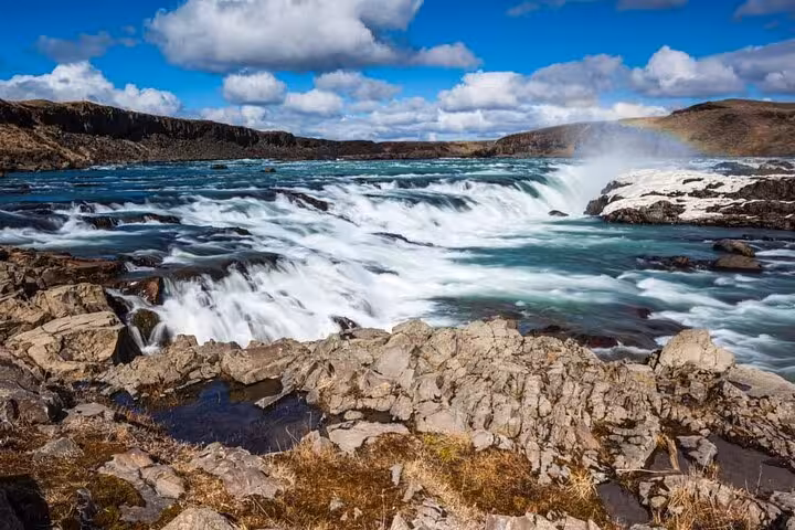Rushing glacial river rapids over rocky terrain under blue sky on Iceland's South Coast