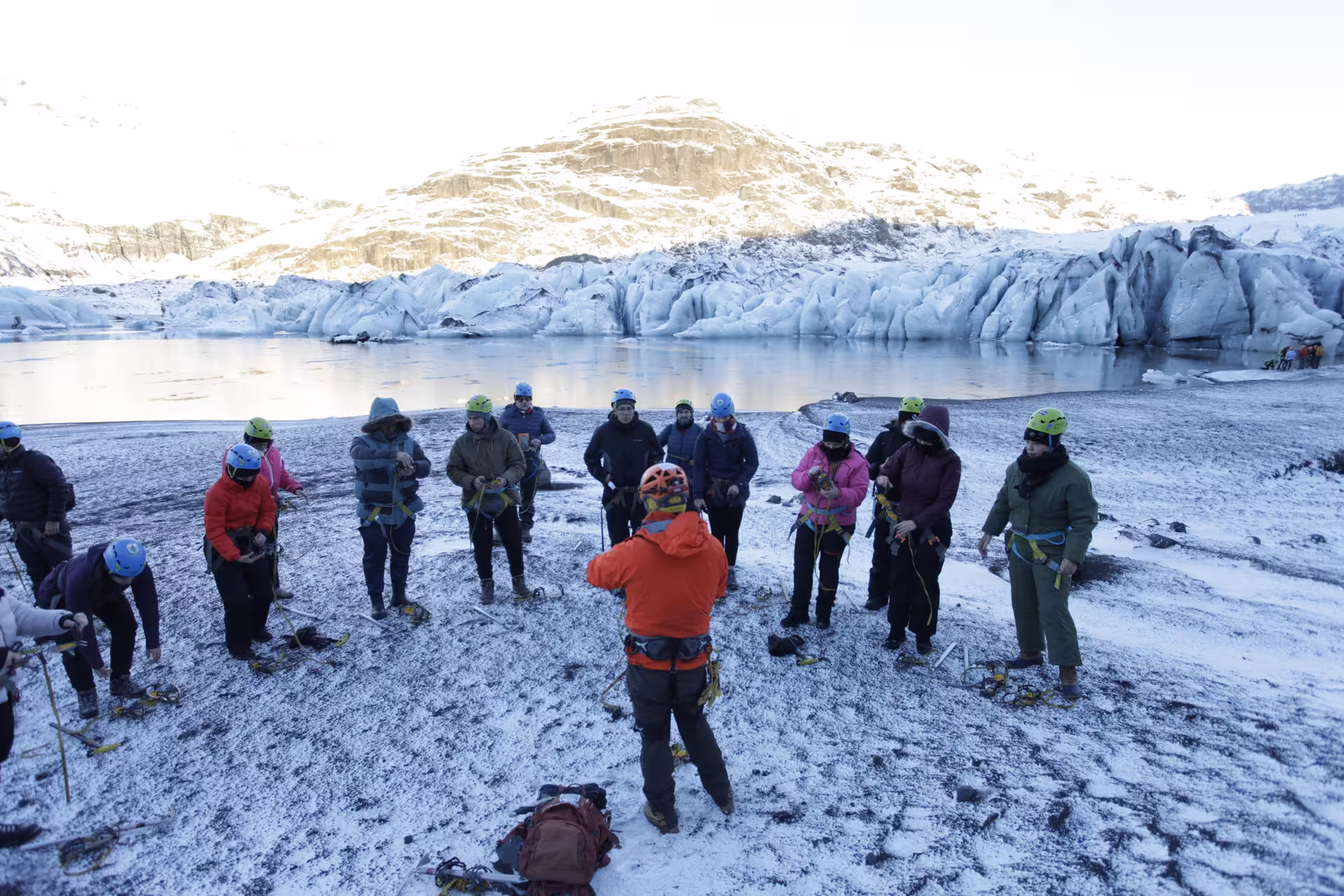 Tour guide briefing group of hikers in helmets and harnesses at Iceland glacier with icy lagoon behind