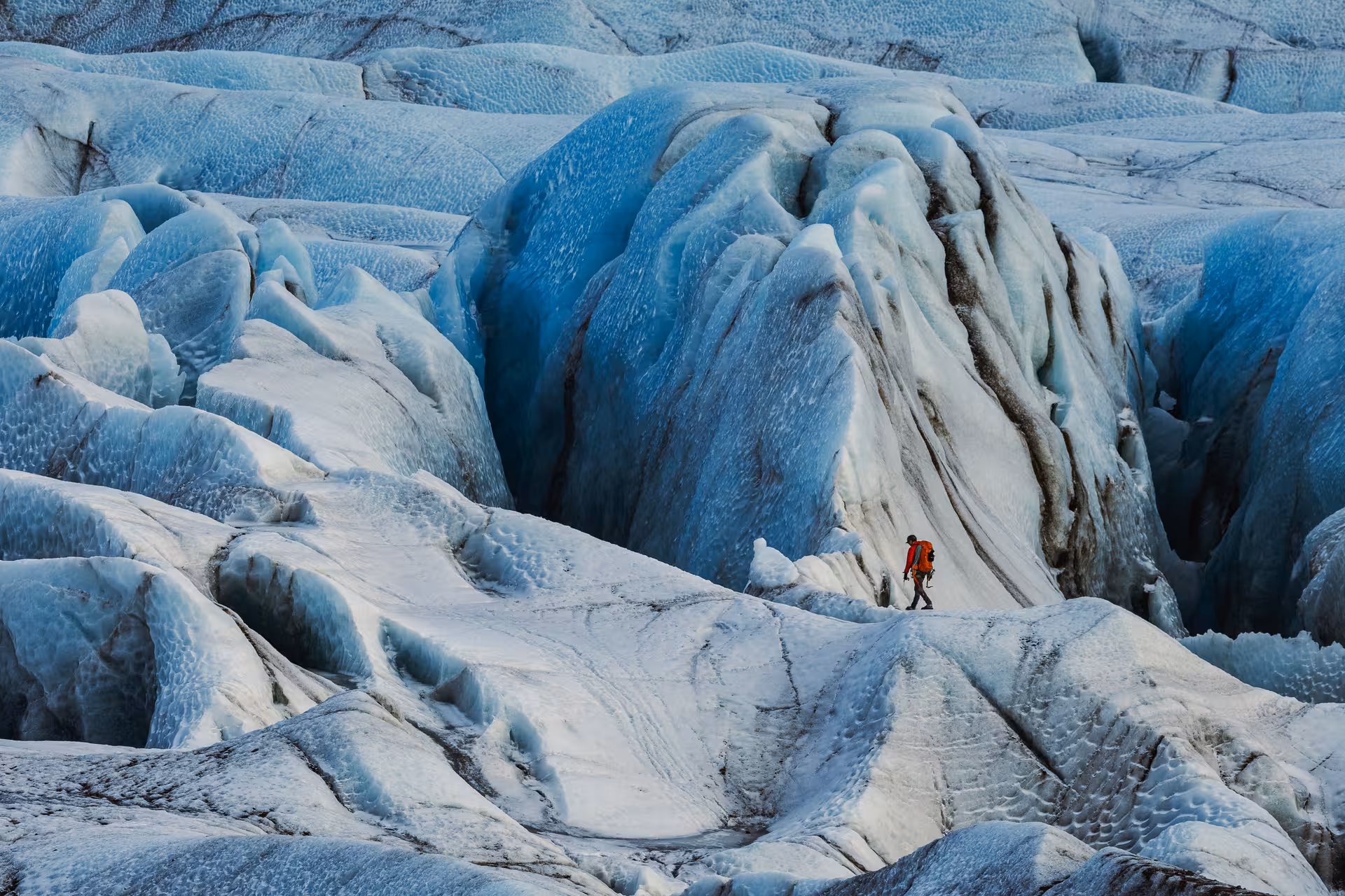 Hiker in orange jacket walking across vast blue glacier ice formations on Iceland's south coast