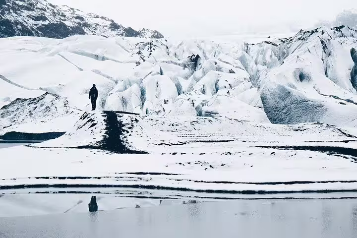 Solo hiker walking on snowy glacier ridge along Iceland's south coast with icy lagoon reflection below