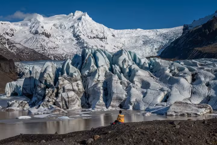 Hiker in yellow jacket sitting before massive glacier with snow-capped mountain on Iceland's south coast
