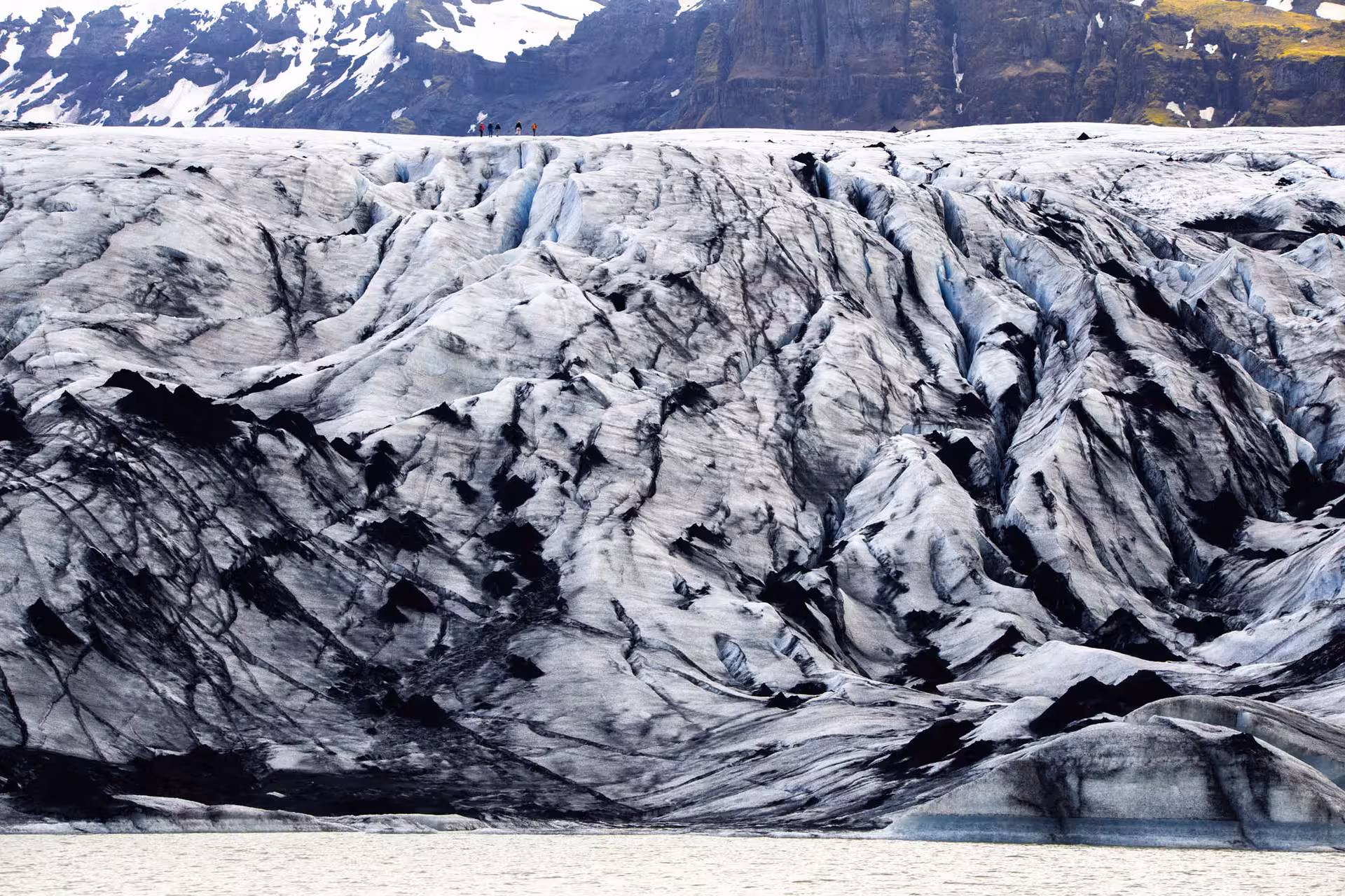 Hikers on a rugged black-streaked glacier with snow-capped mountains on Iceland's South Coast