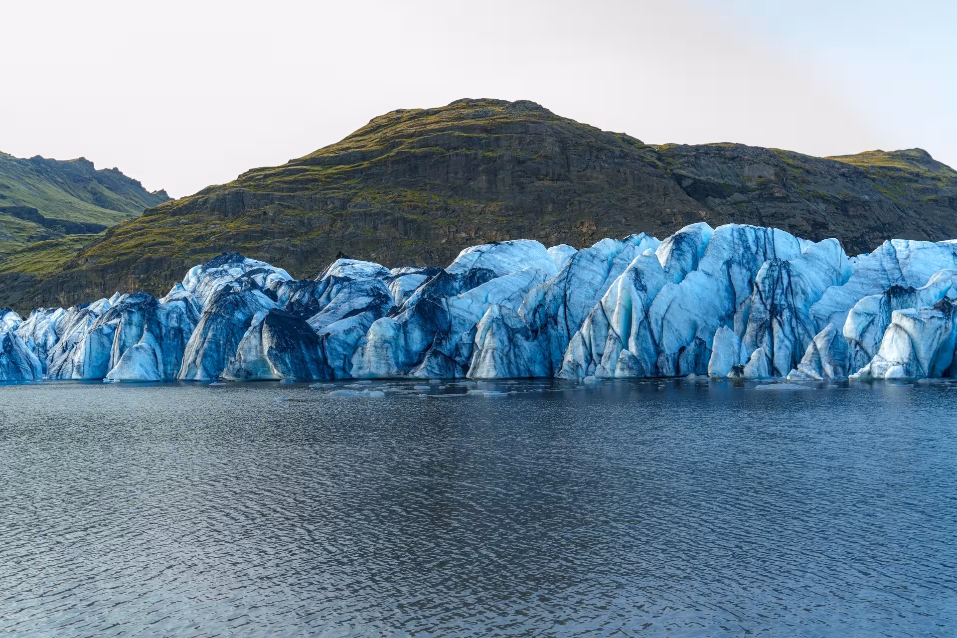 Blue glacier ice formations meeting glacial lagoon with green volcanic mountains in Iceland's south coast