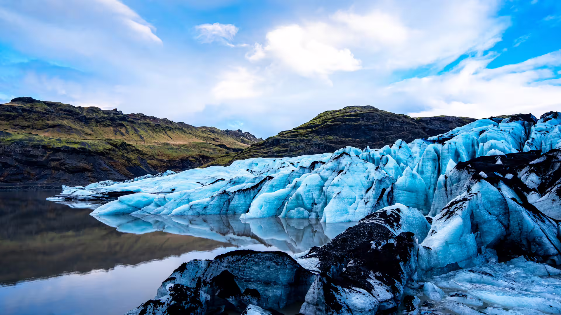 Blue glacial ice formations reflecting in a still lagoon with green volcanic hills in Iceland