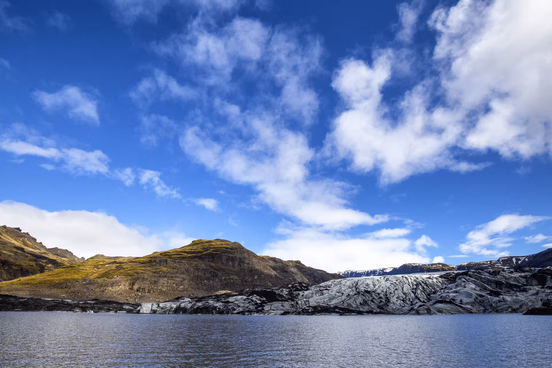 Glacier tongue meeting a glacial lagoon with moss-covered volcanic mountains under blue sky in Iceland