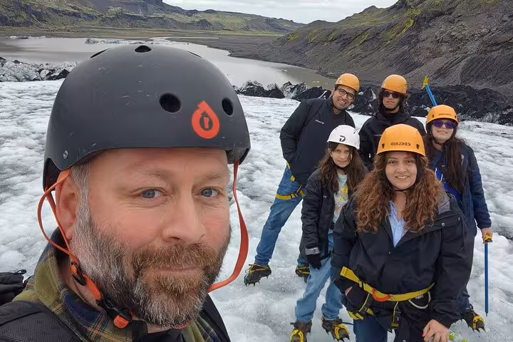 Group of tourists wearing helmets and crampons posing for selfie on Iceland glacier with volcanic mountains behind