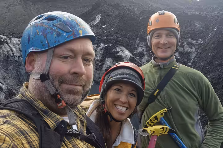 Three smiling hikers wearing climbing helmets pose for selfie on Iceland glacier tour