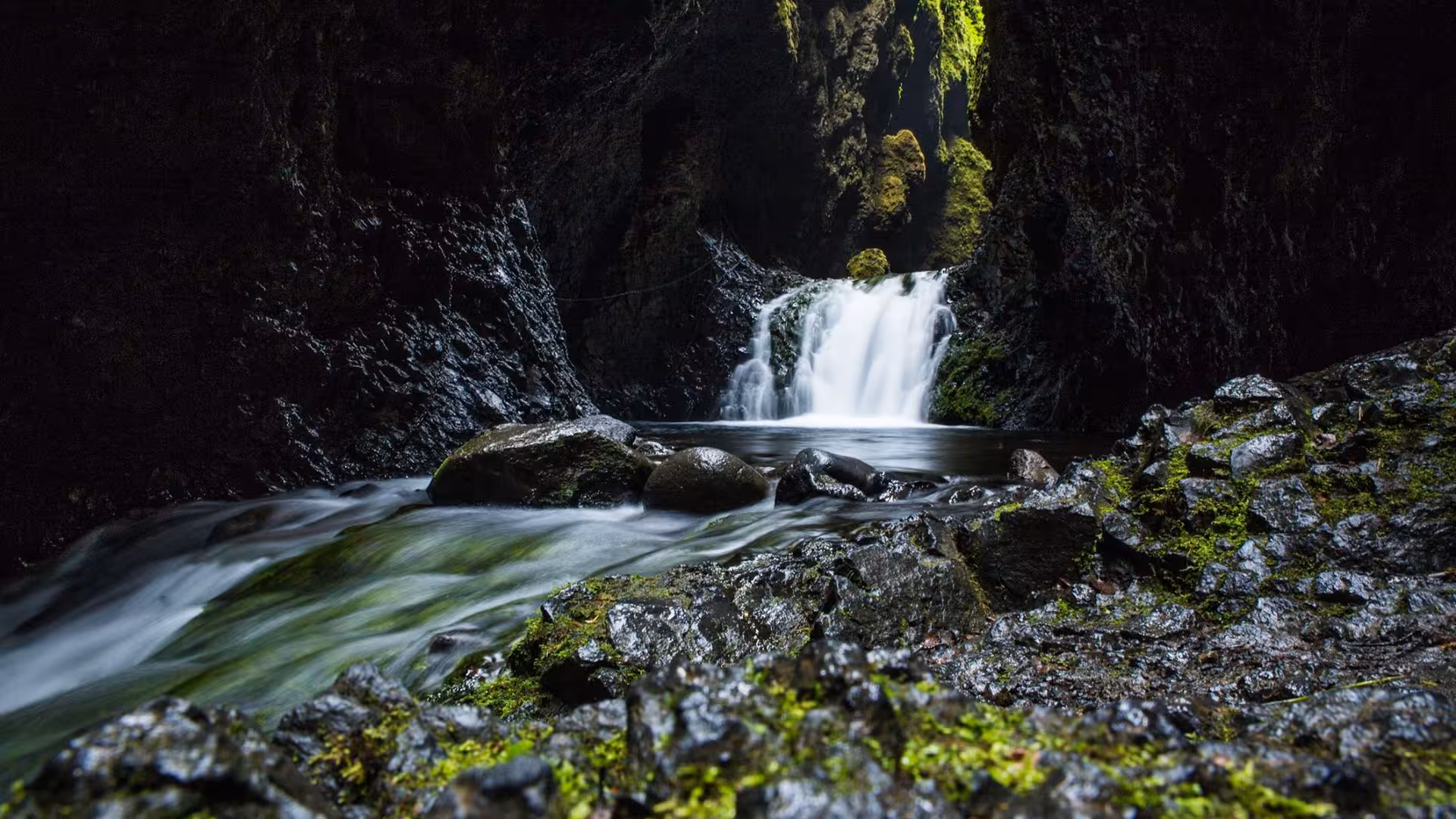 Hidden waterfall cascading into a dark mossy canyon with smooth flowing stream in Iceland