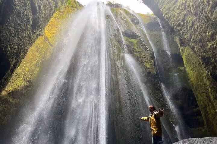 Traveler in yellow jacket standing before cascading waterfall inside mossy canyon on Iceland South Coast