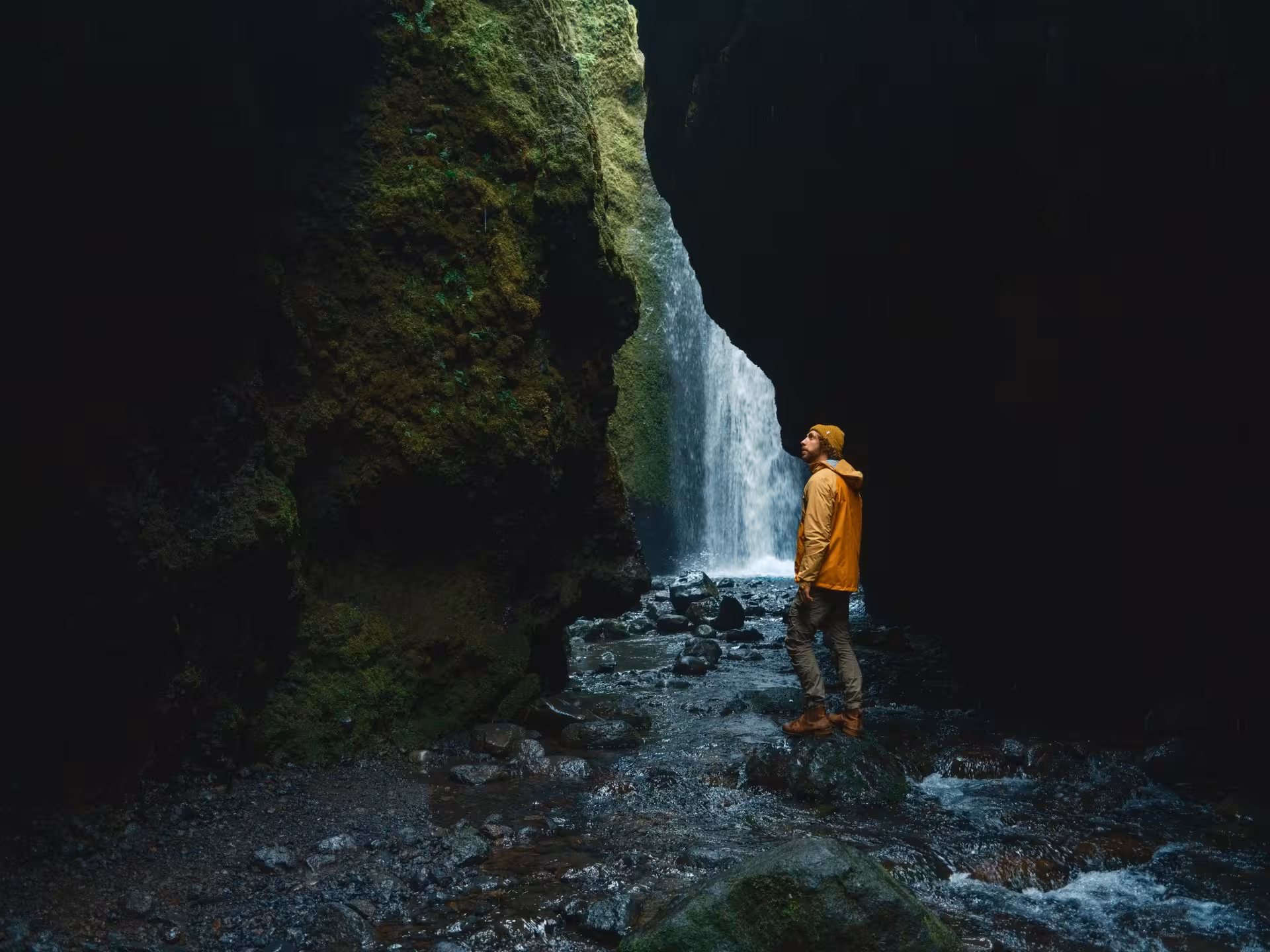 Hiker in yellow jacket standing before a hidden waterfall in a dark mossy canyon on Iceland's south coast