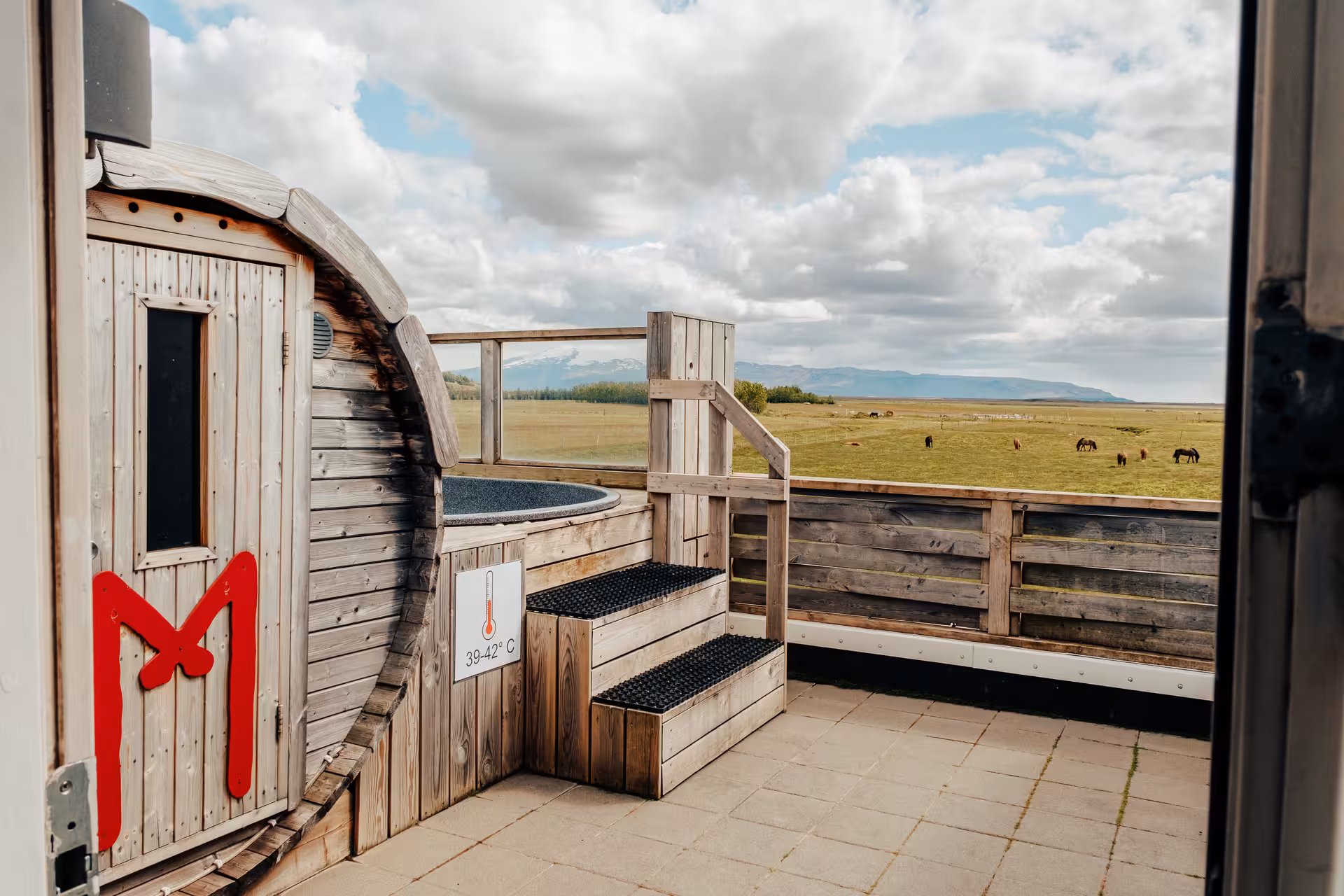 Outdoor hot tub at 39-42°C on wooden deck overlooking Icelandic green fields with grazing horses and mountains