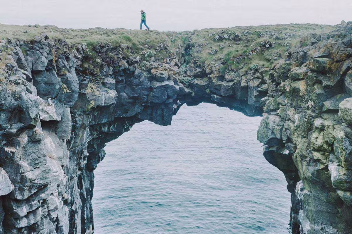 Hiker walking across a natural rock arch bridge over the ocean on Iceland's South Coast