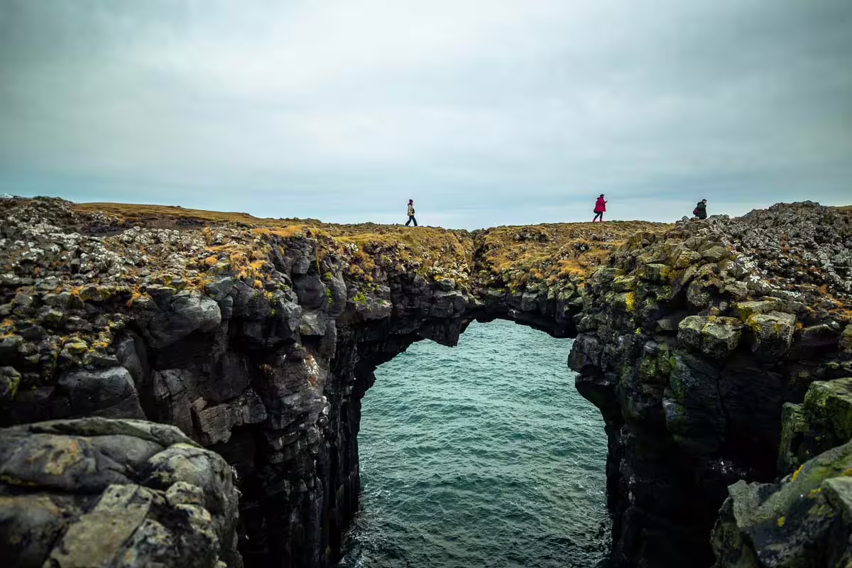 Hikers crossing a natural rock arch bridge over turquoise ocean waters on Iceland's rugged coastline