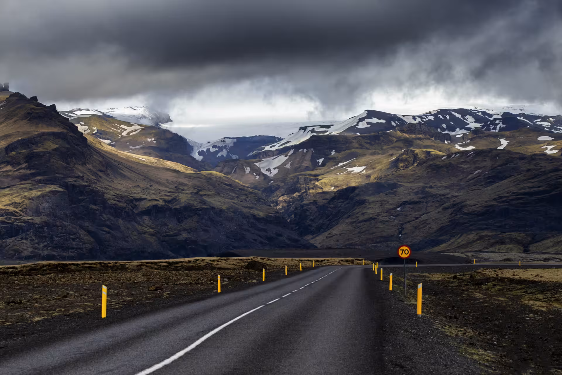 Two-lane road leading toward snow-capped mountains under stormy skies on Iceland's South Coast