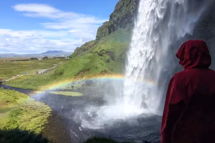 Person in red hooded jacket viewing Seljalandsfoss waterfall with rainbow on Iceland's South Coast