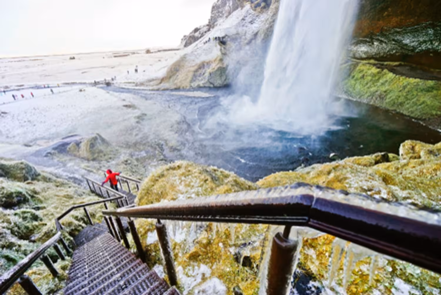 Visitor in red jacket on icy staircase beside Seljalandsfoss waterfall on Iceland South Coast winter tour