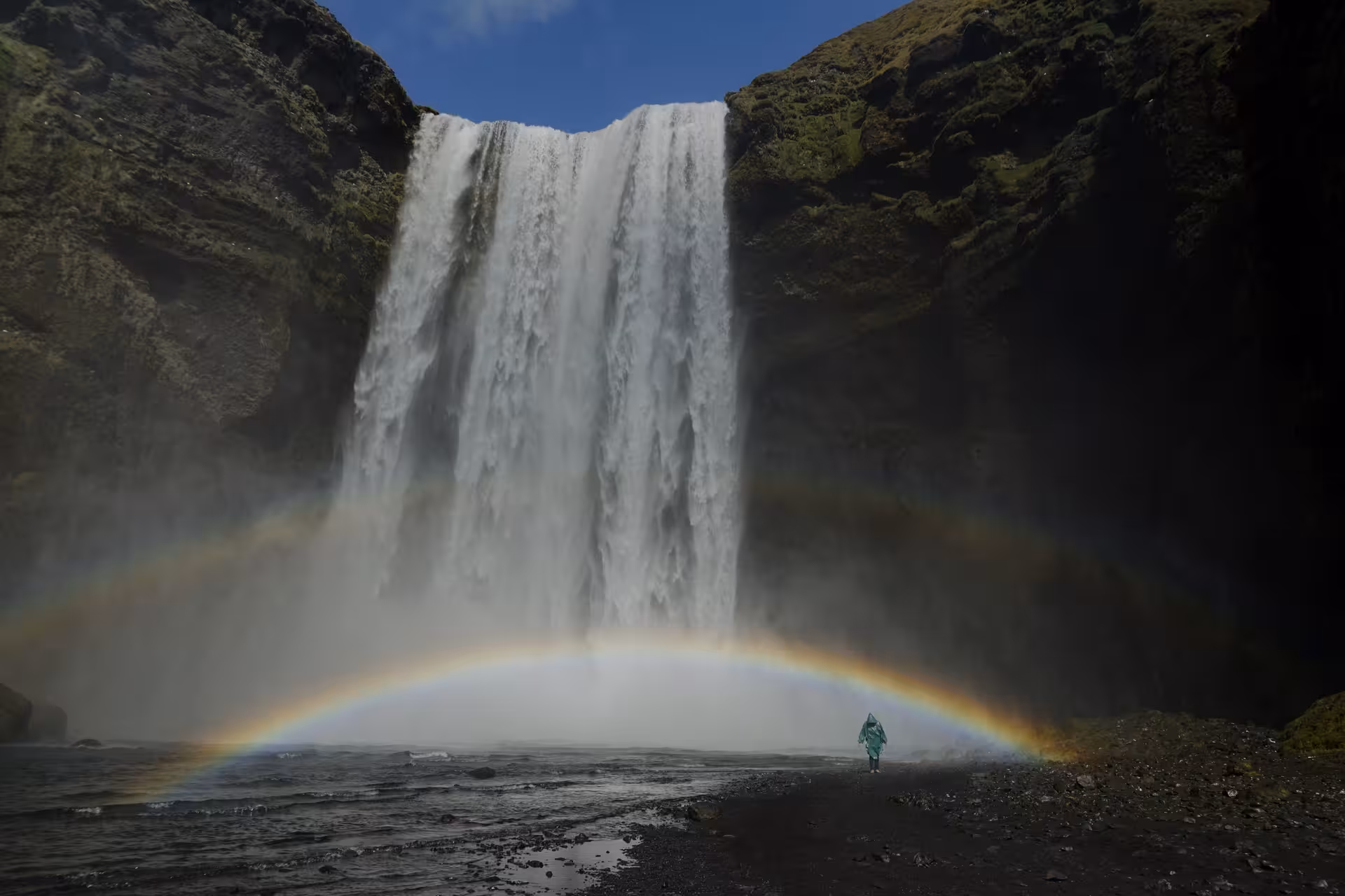 Person in green raincoat standing before Skógafoss waterfall with a full rainbow arc in Iceland