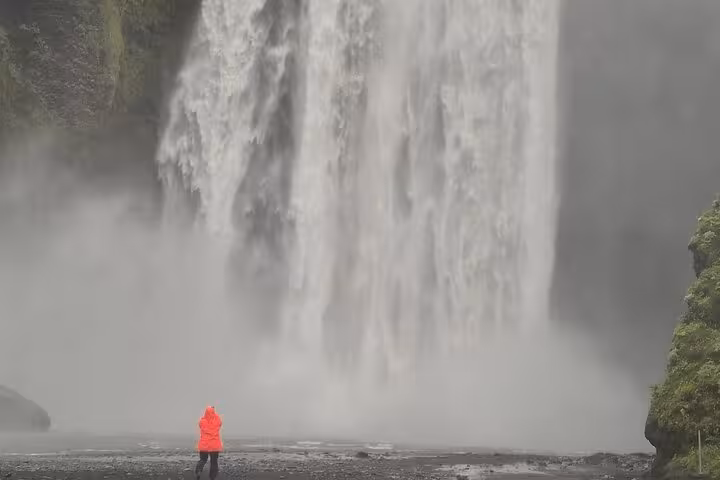 Person in red jacket standing before massive Skógafoss waterfall on Iceland's South Coast