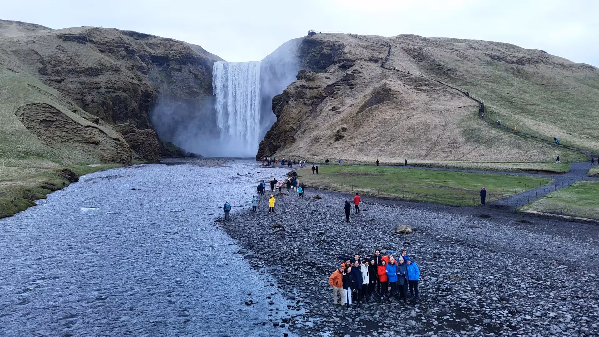 Tour group posing at Skógafoss waterfall on Iceland's South Coast with rocky riverbed and misty cascade