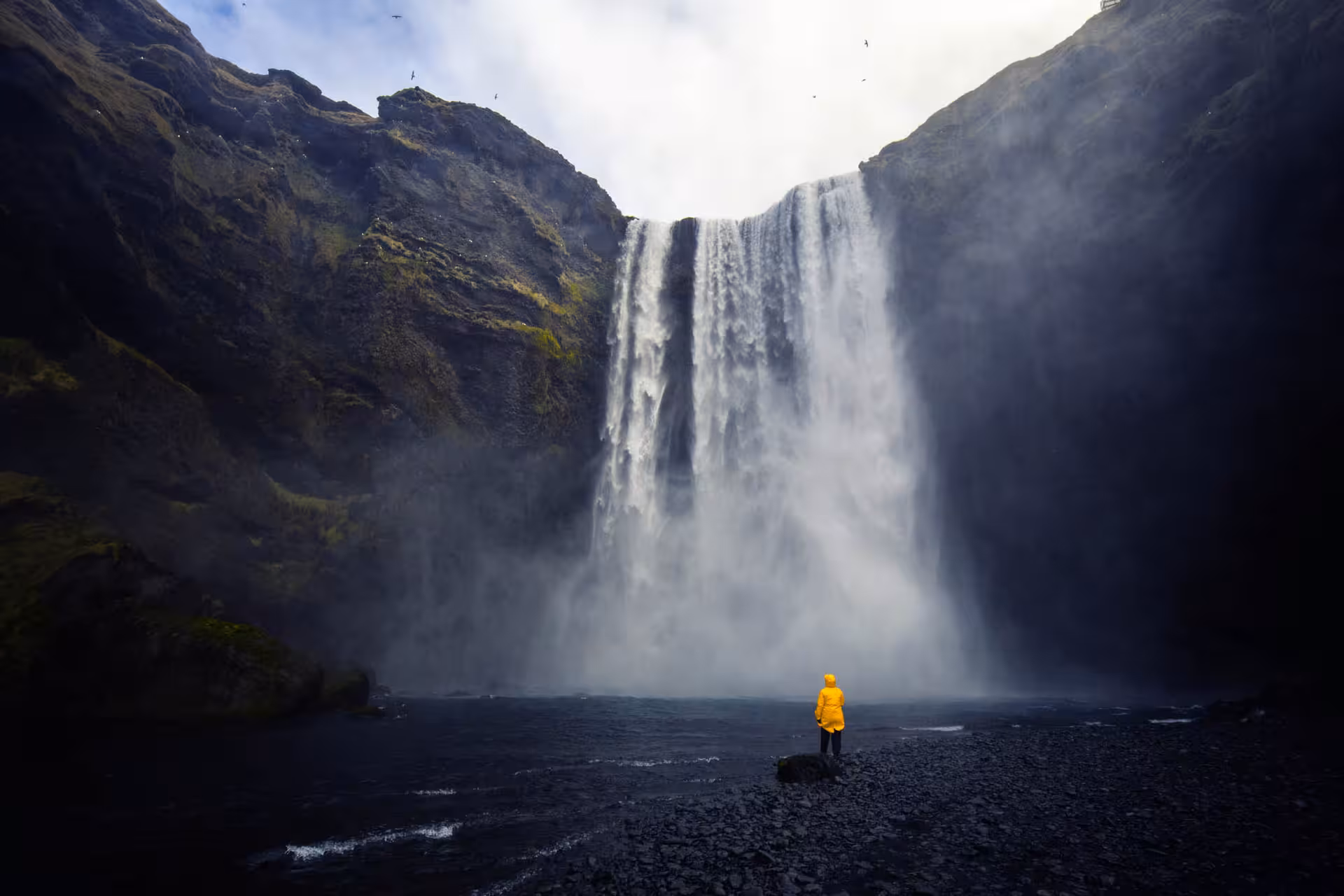 Person in yellow jacket standing before massive Skógafoss waterfall on Iceland's south coast
