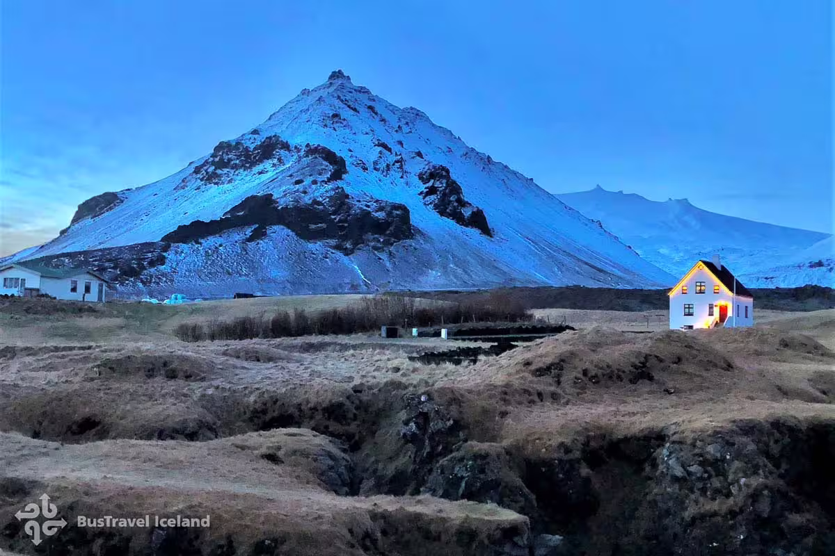 Snow-capped volcanic mountain with illuminated white farmhouse at dusk on Iceland's South Coast