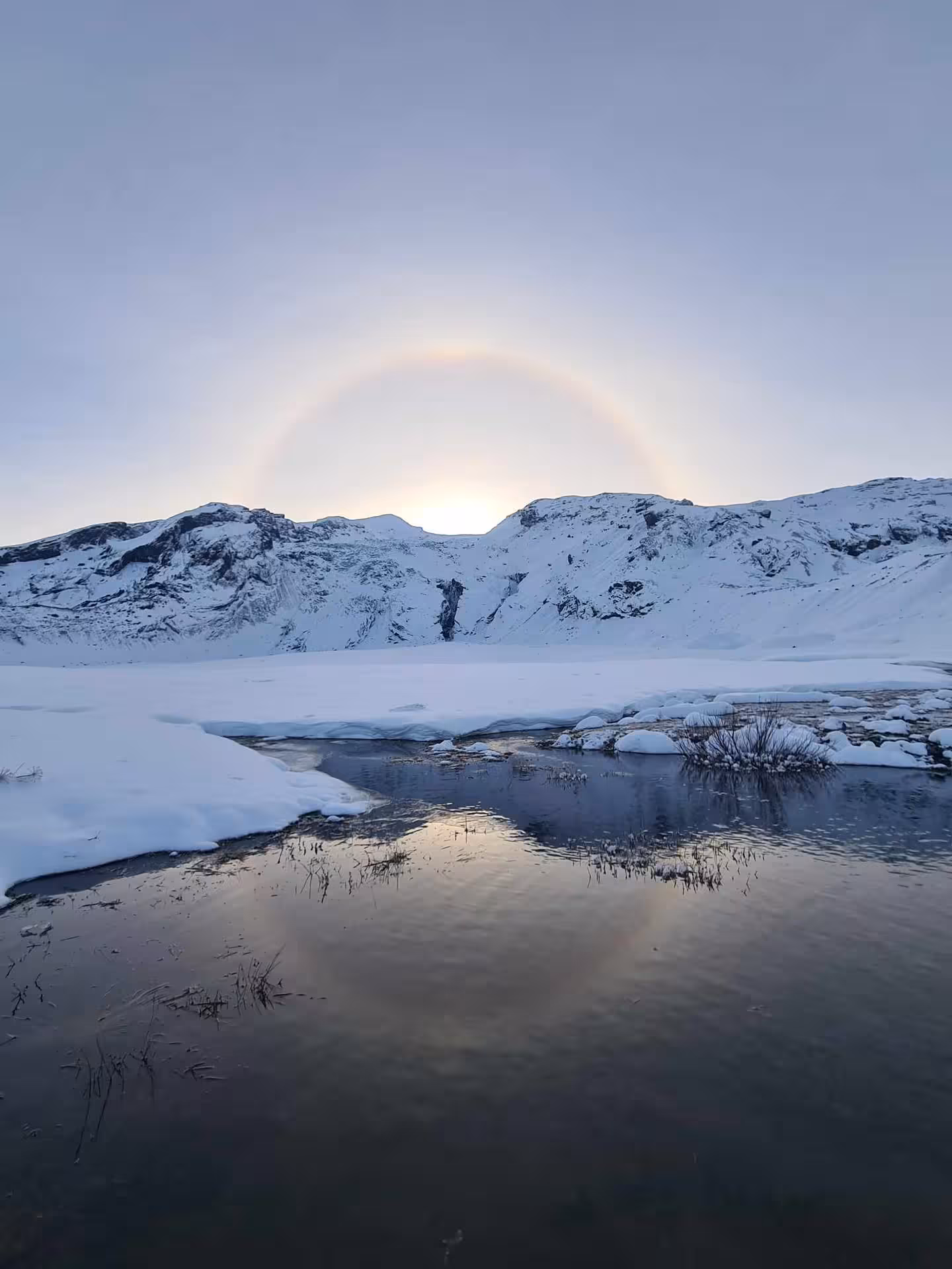 Solar halo over snow-covered mountains reflected in a partially frozen lake on Iceland's South Coast