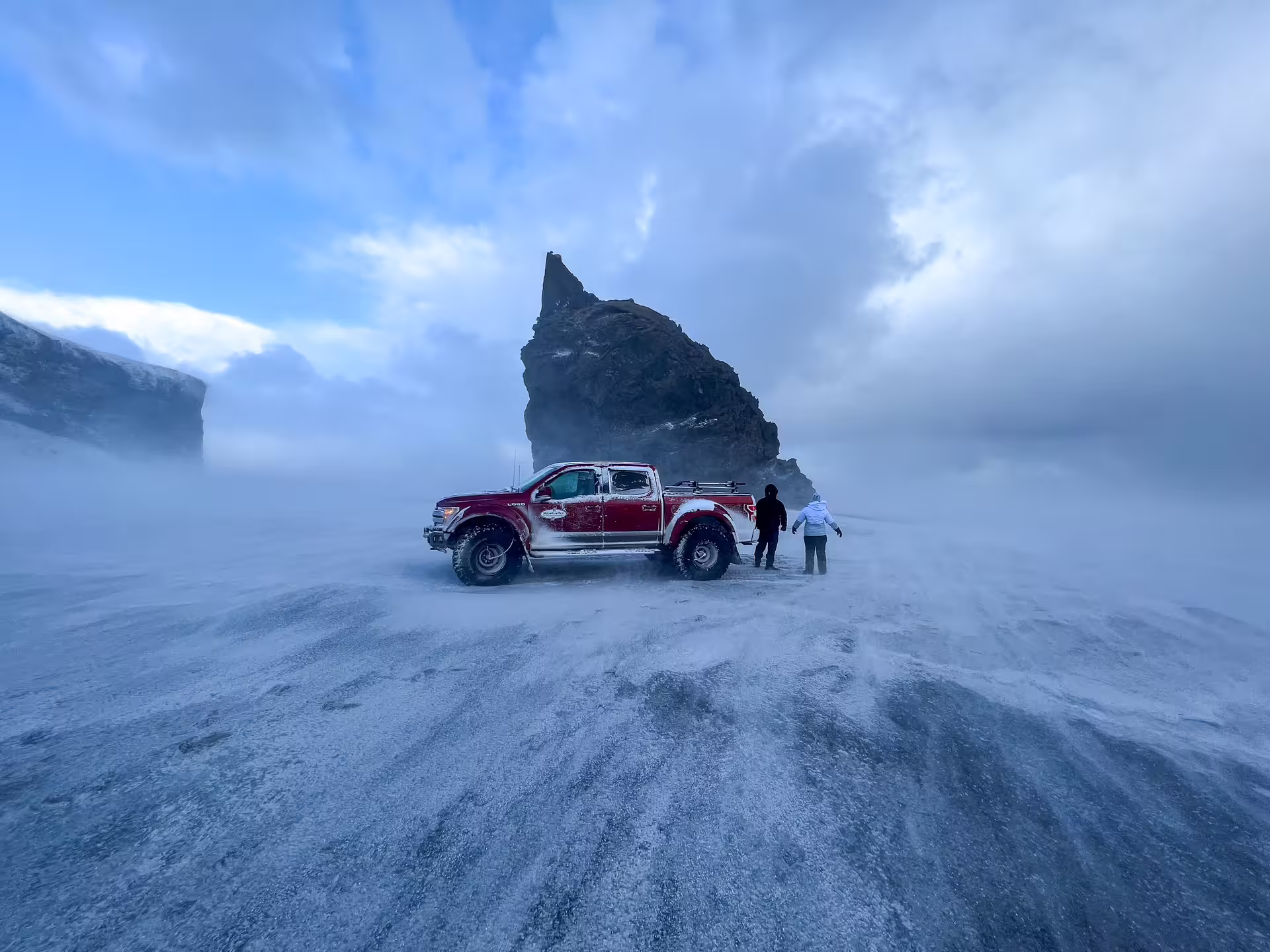 Red super jeep and two people beside a volcanic rock spire on Iceland's snow-swept south coast
