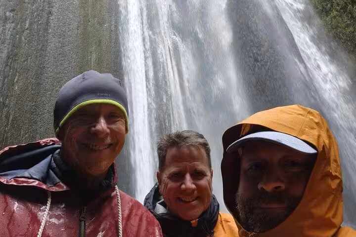 Three smiling men in rain gear posing in front of a large waterfall on Iceland's South Coast