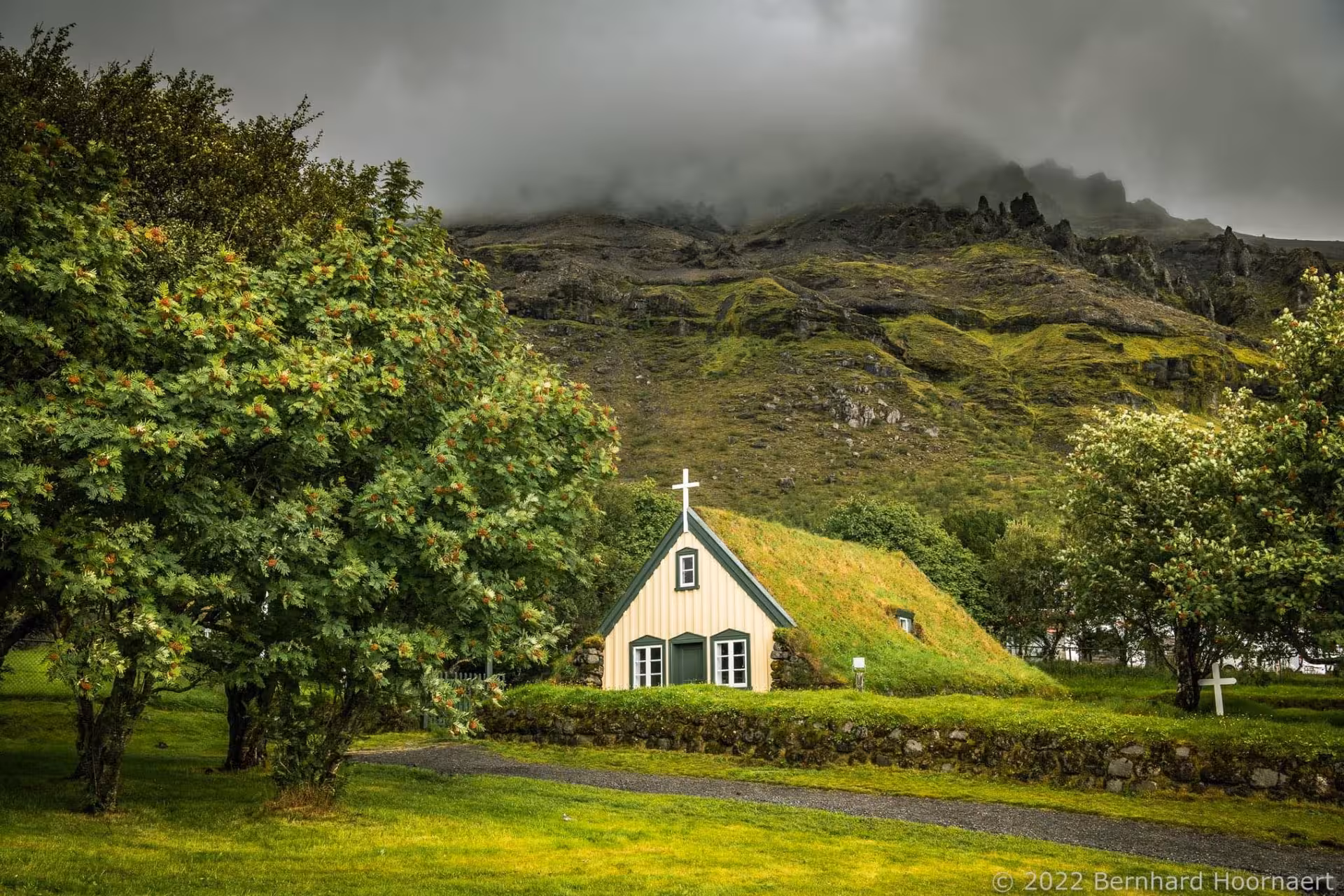 Traditional Icelandic turf church with grass roof surrounded by trees and dramatic mountain backdrop on Iceland's South Coast