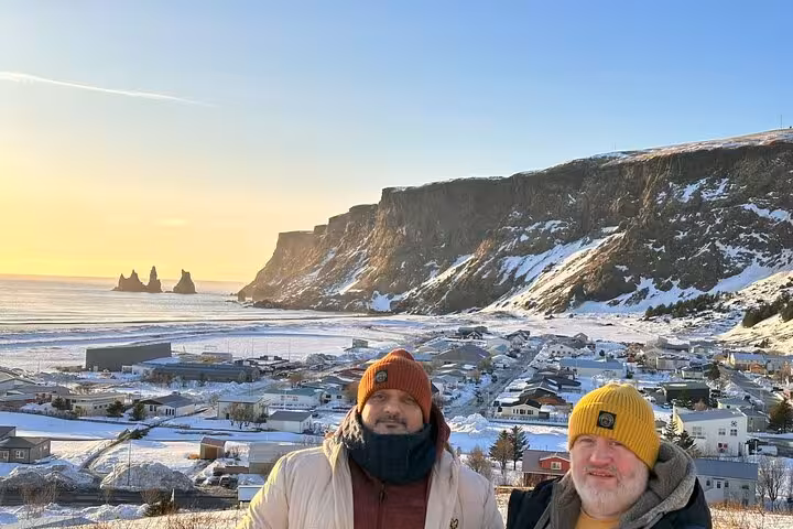Two travelers posing above snowy Vík village with Reynisdrangar sea stacks and cliffs on Iceland's South Coast