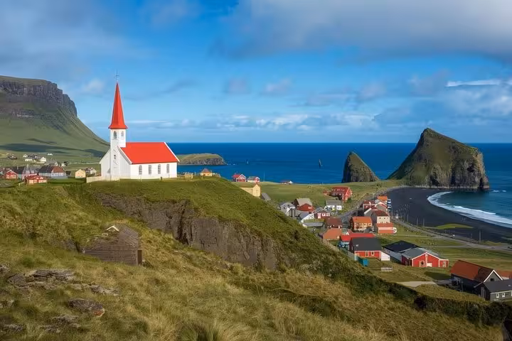White church with red roof overlooking colorful village, black sand beach, and dramatic sea stacks on Iceland's south coast