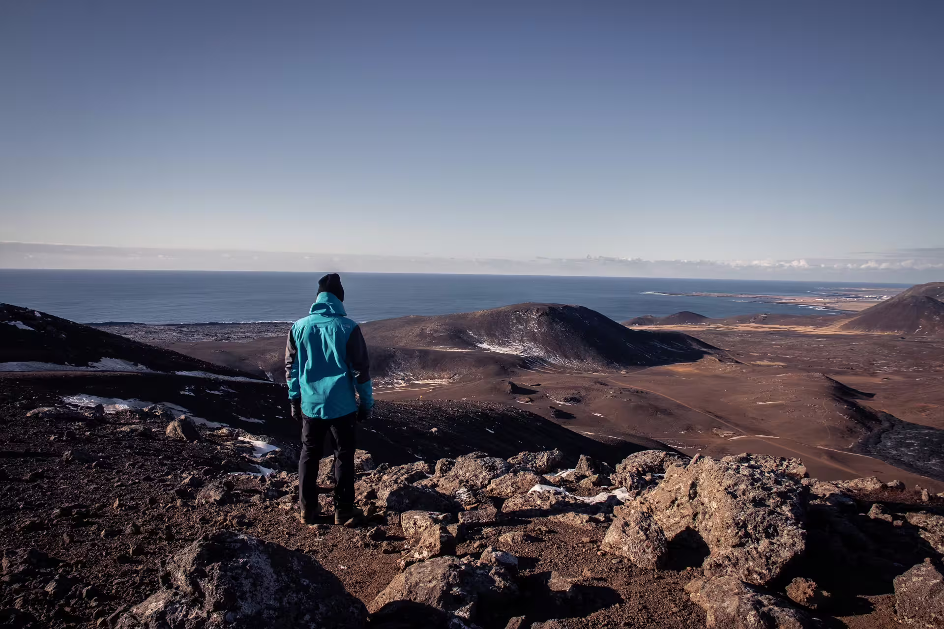 Hiker in teal jacket standing on volcanic ridge overlooking Iceland's rugged lava fields and coastline