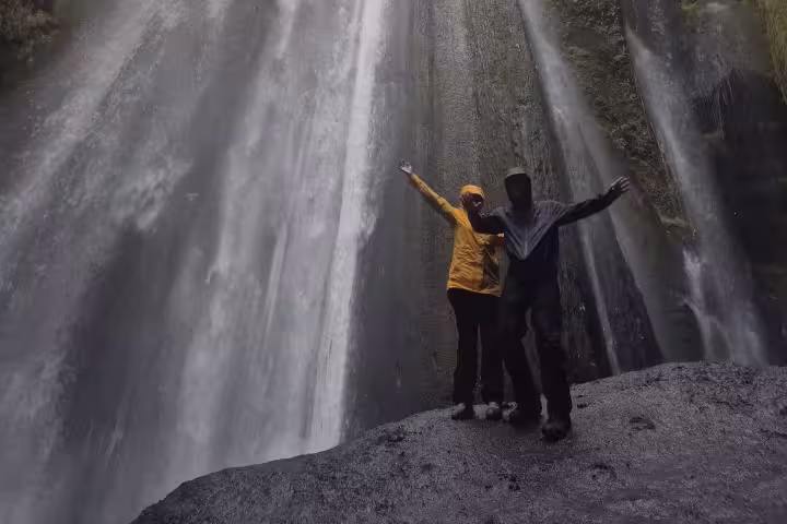 Two travelers with arms raised standing before a towering waterfall in a mossy canyon on Iceland's South Coast