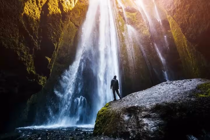Person standing on rock gazing at massive waterfall inside a mossy canyon cave in Iceland