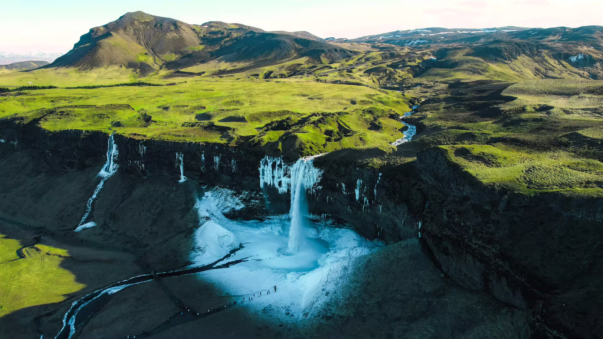 Aerial view of Seljalandsfoss waterfall surrounded by frozen basin and lush green Icelandic highlands