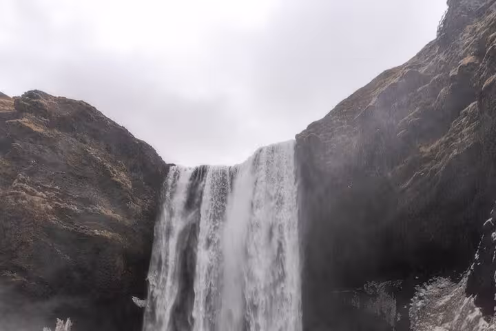 Powerful waterfall cascading between dark rocky cliffs on Iceland's south coast under overcast skies