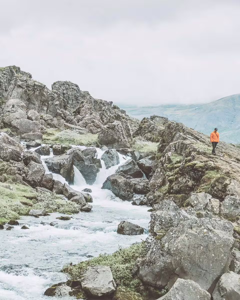 Hiker in orange jacket standing beside rocky waterfall on Iceland's South Coast mountain trail