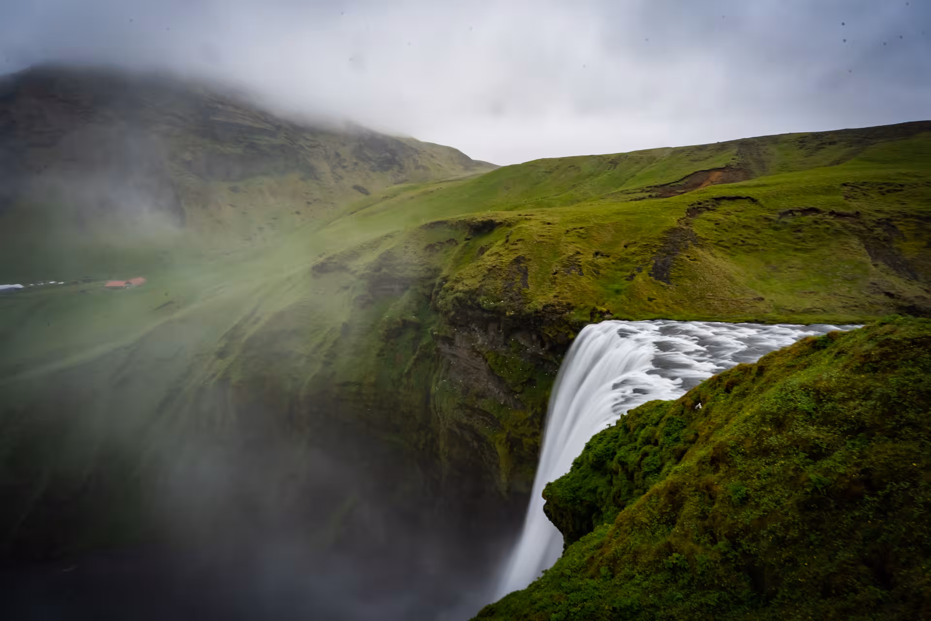 Skógafoss waterfall cascading over mossy cliffs with mist and green hills on Iceland's South Coast
