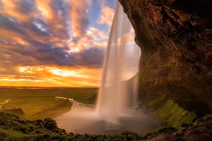 View from behind Seljalandsfoss waterfall in Iceland at golden sunset with green plains beyond