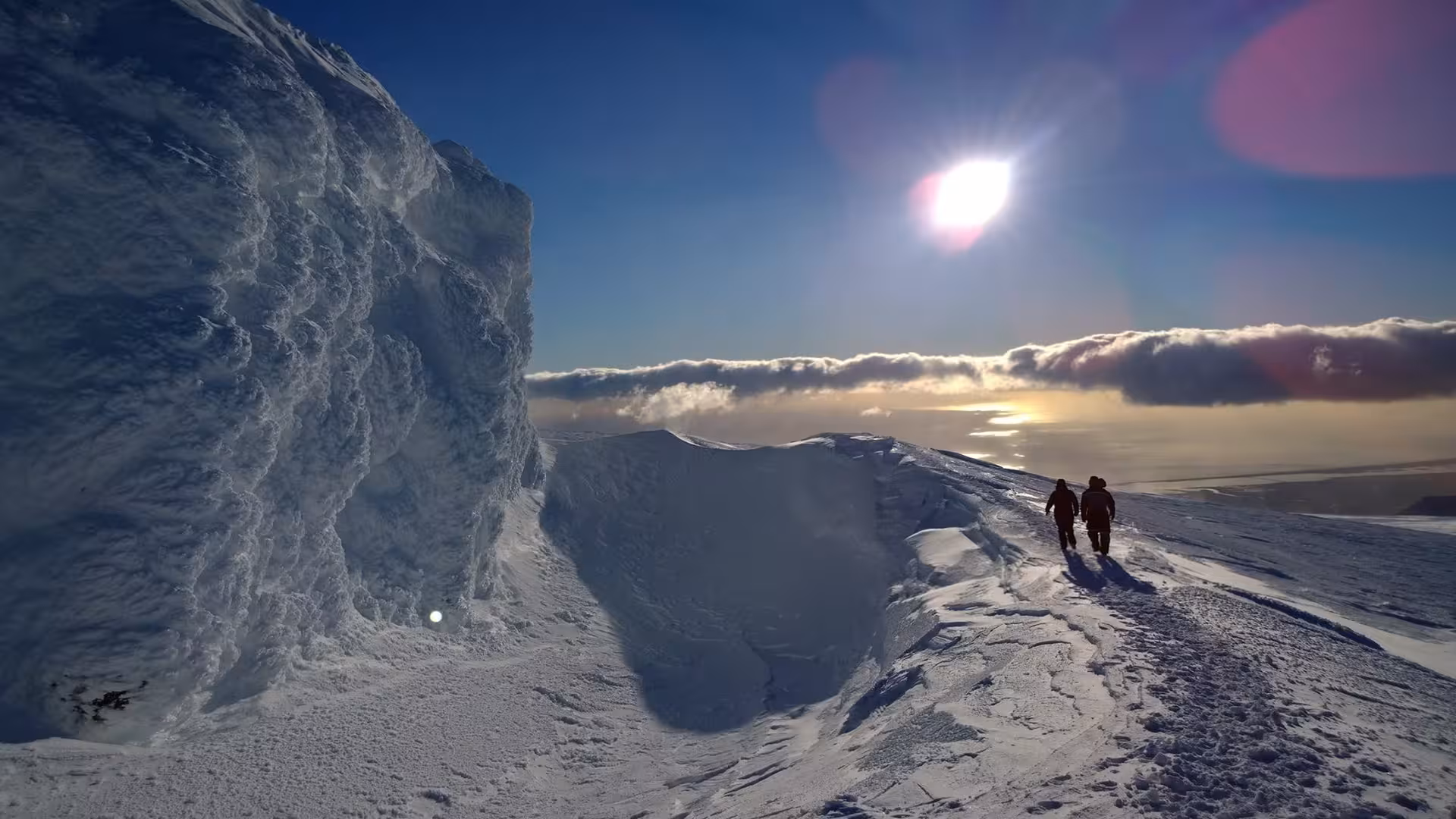 Two hikers walking along a snowy mountain ridge in Iceland with bright sun and clouds below