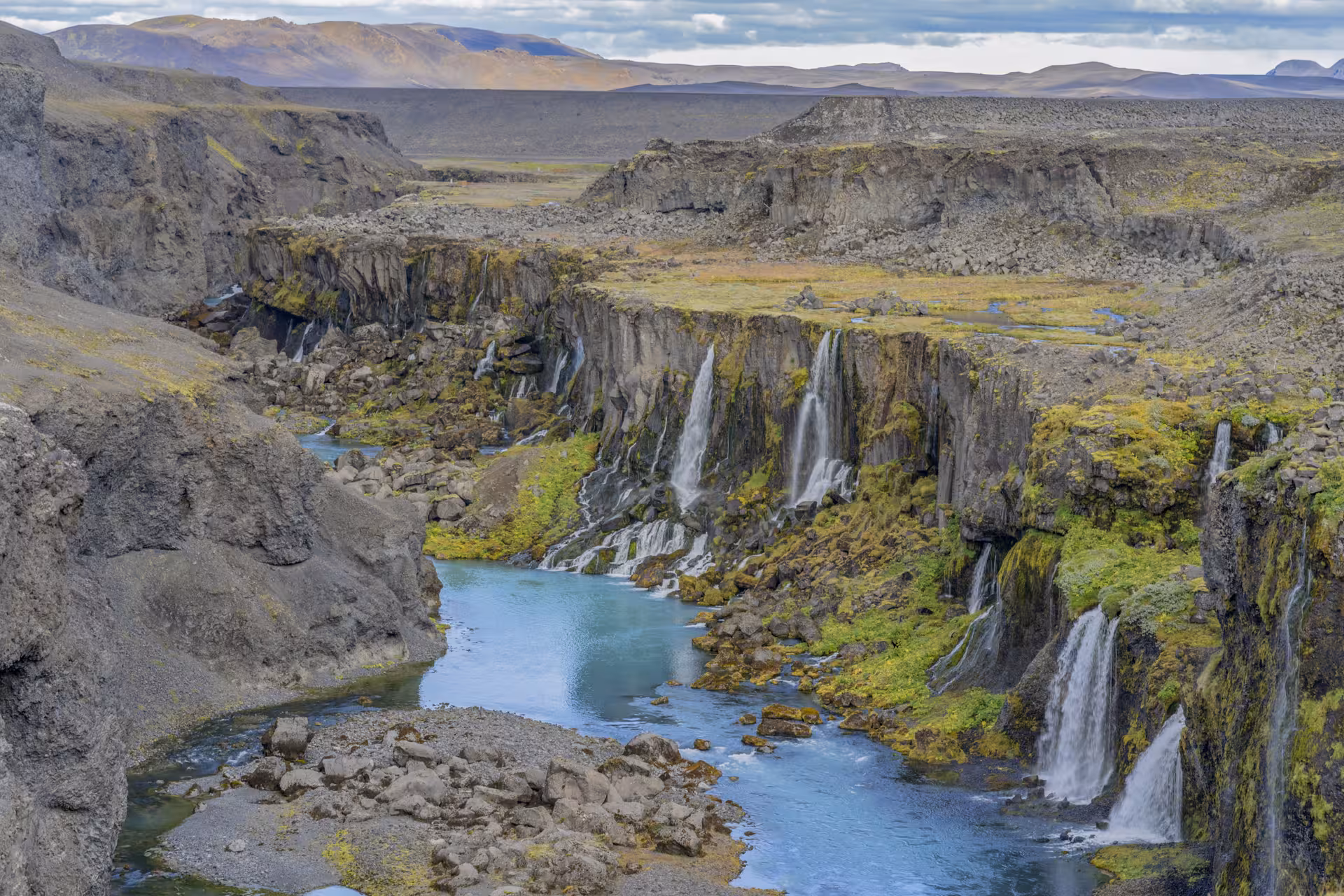 Multiple waterfalls cascading into a turquoise river canyon with moss-covered basalt cliffs in Iceland