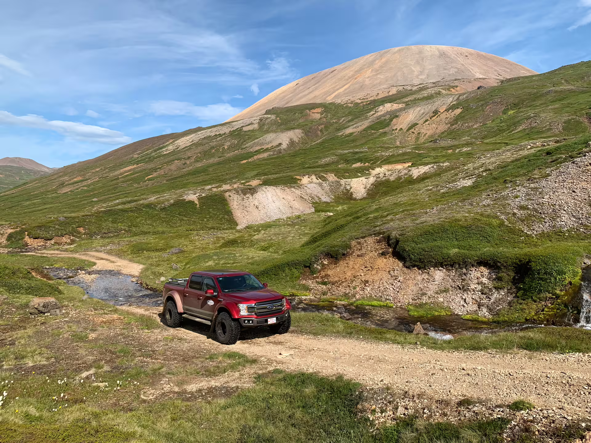 Red lifted Ford F-150 super jeep on a dirt trail with a green volcanic mountain in Iceland