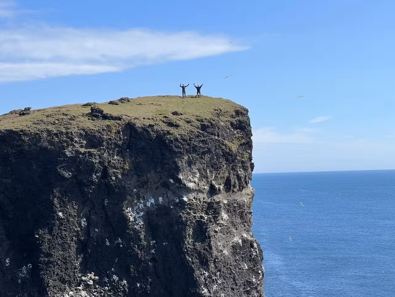 Two people with arms raised on top of dramatic sea cliffs in Iceland on a sunny day