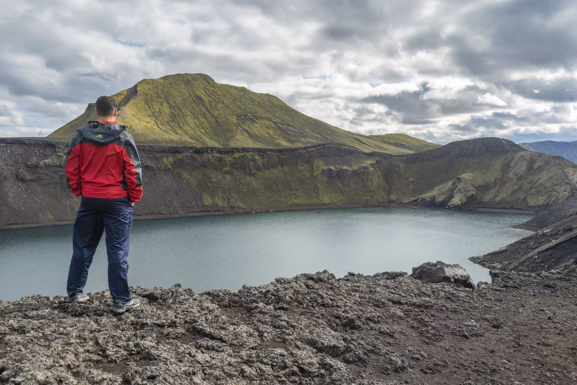 Hiker in red jacket overlooking volcanic crater lake with green moss-covered mountains in Iceland