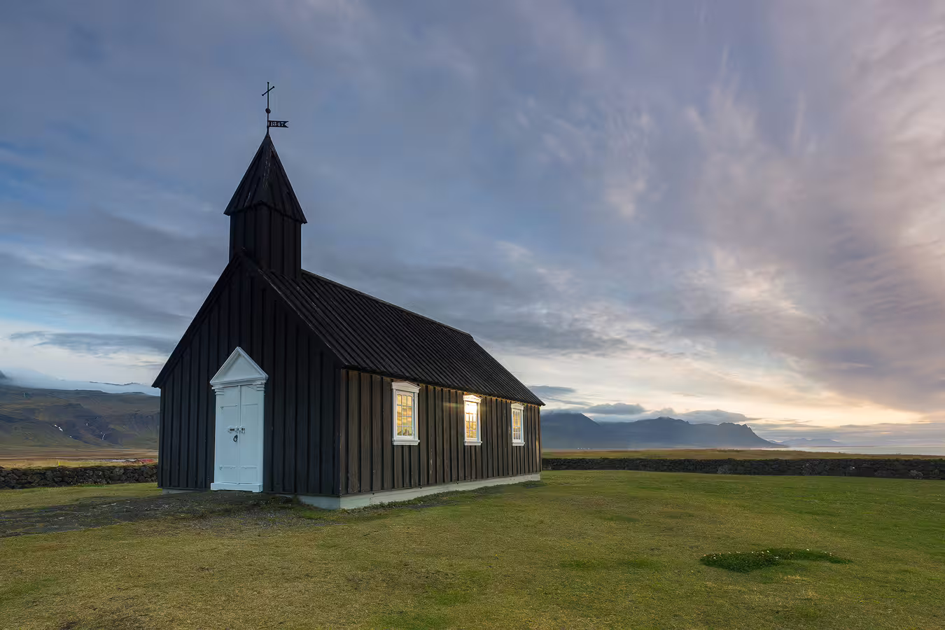 Black wooden church with white door and glowing windows on green Icelandic landscape at dusk