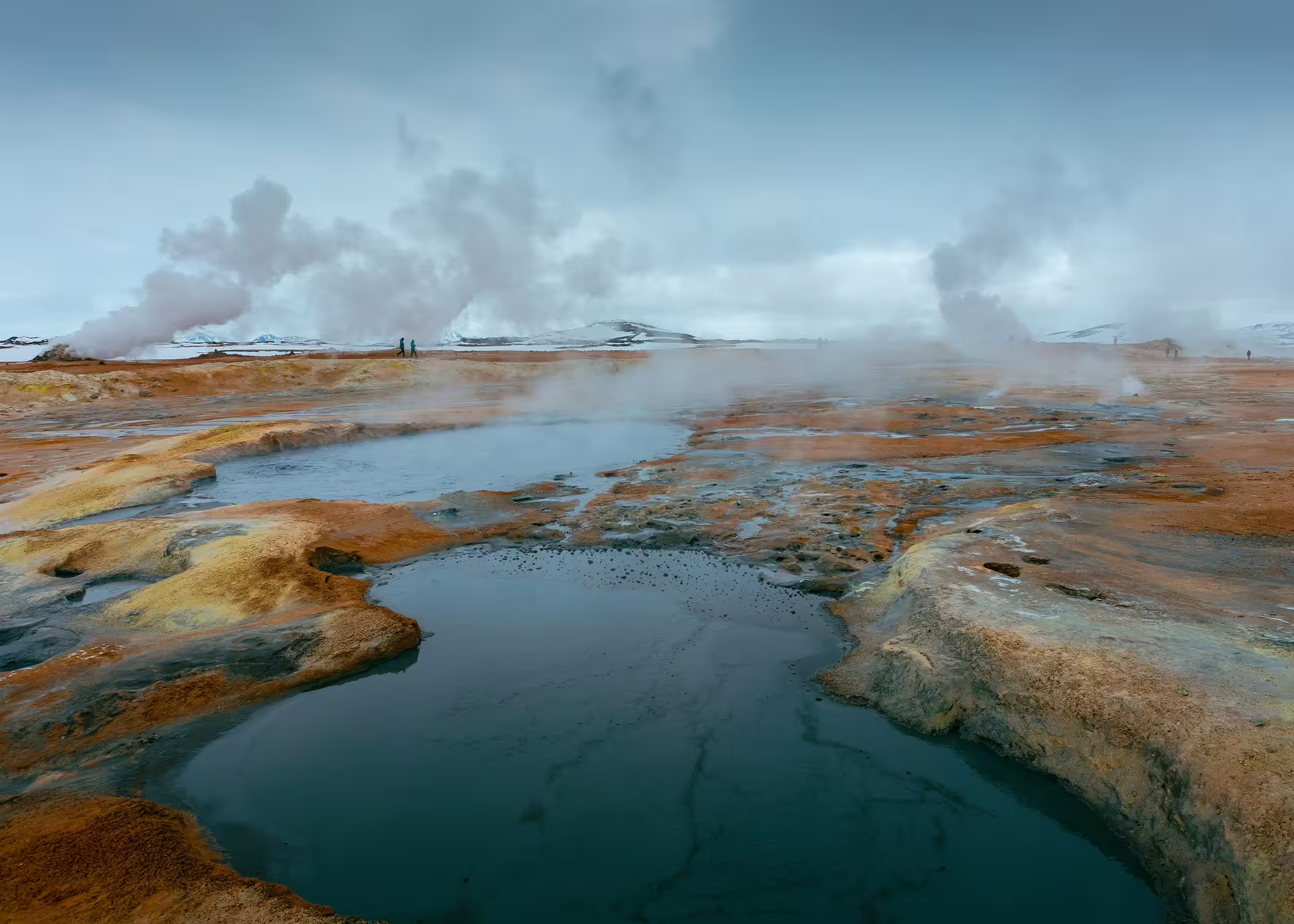 Steaming geothermal hot springs with orange mineral deposits and snow-capped mountains in Iceland